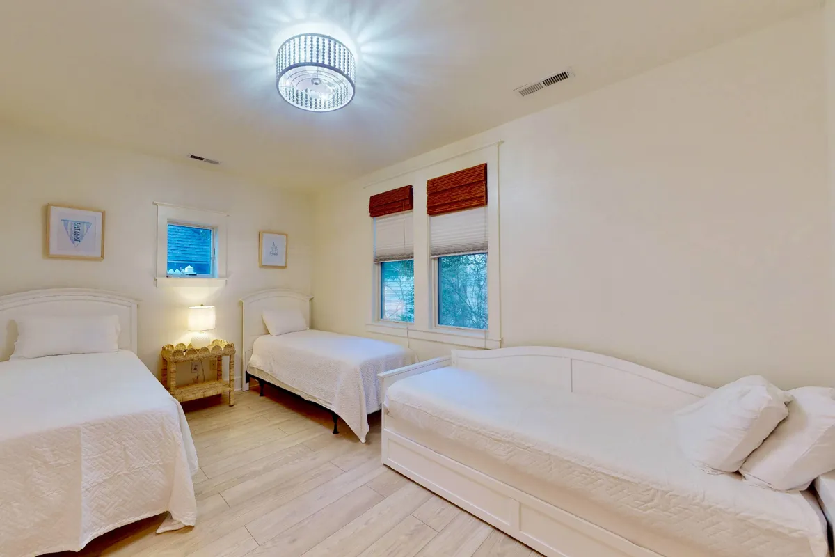 A cozy bunk room with three beds, a distinctive chandelier light fixture, and windows with blinds. The space is inviting and versatile, suitable for children or additional guests seeking comfort and flexibility.