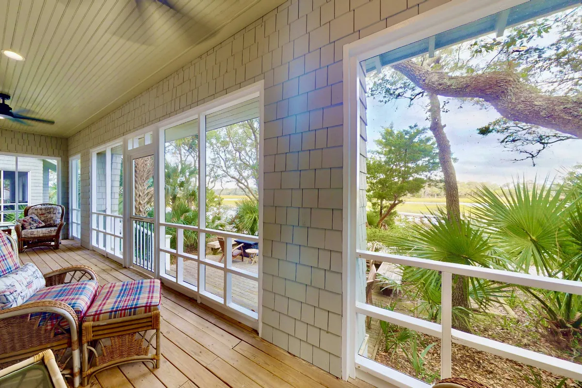 A bright living area with woven chairs and expansive large windows that frame lush natural greenery and marsh views. The space features wooden flooring and is bathed in natural sunlight, with a peaceful atmosphere ideal for relaxation or morning coffee.