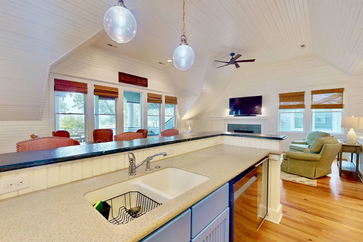 A modern kitchen with a large island and sink, seamlessly connected to a comfortable seating area. The space is lined with large windows that let in marsh views and sunlight, fostering a welcoming, open environment ideal for both cooking and socializing.