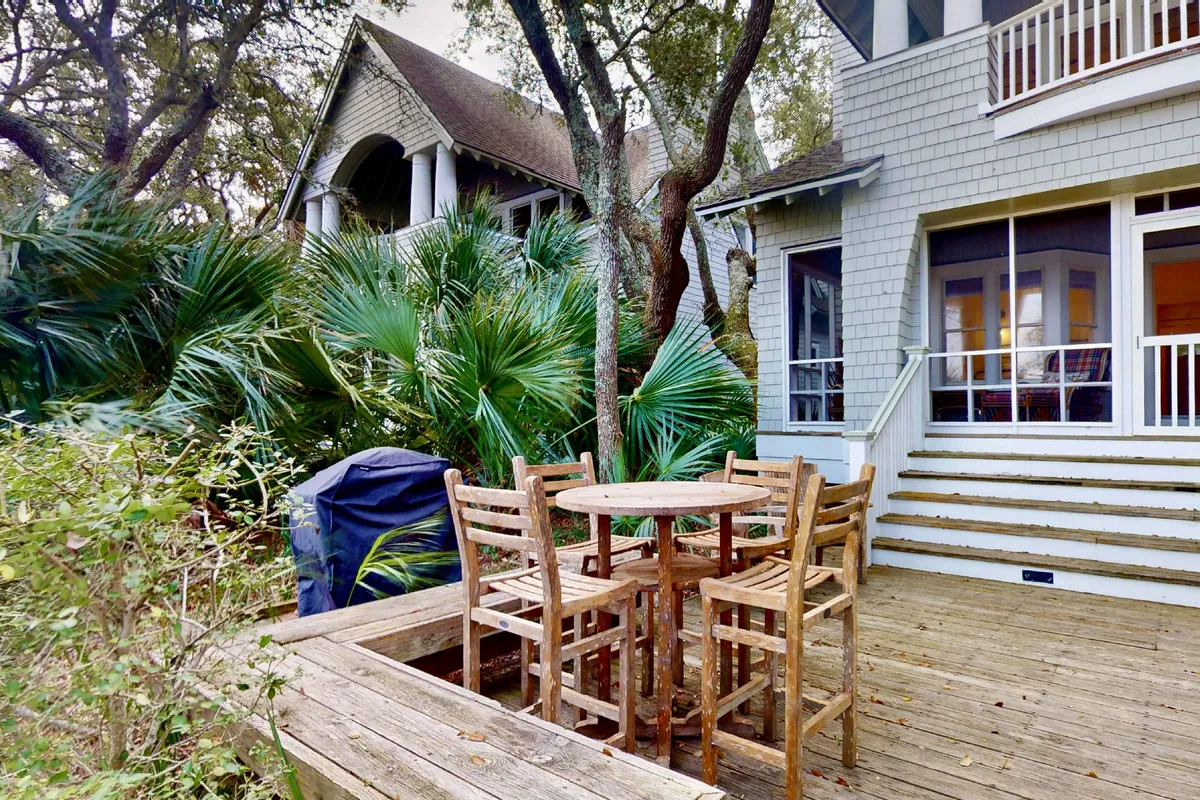 This image features a wooden outdoor dining table with stool seating, surrounded by lush greenery and adjacent to the house. The setup is perfect for gatherings, al fresco meals, and evening relaxation under the stars, showcasing a central space for socializing and entertaining.