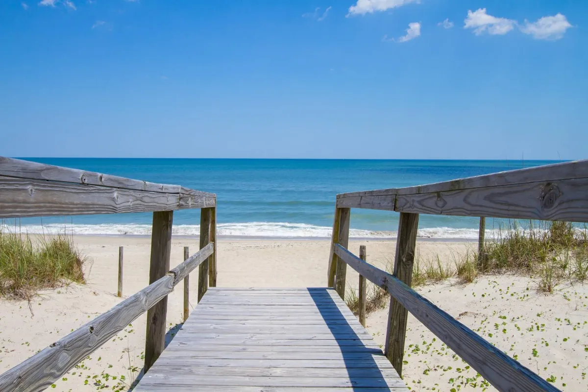 A wooden pathway leads across the sand dunes to the expansive ocean, framed by blue skies. The scene invites families to enjoy beach walks, sandcastle building, swimming, and picnicking—activities highlighted in the section.