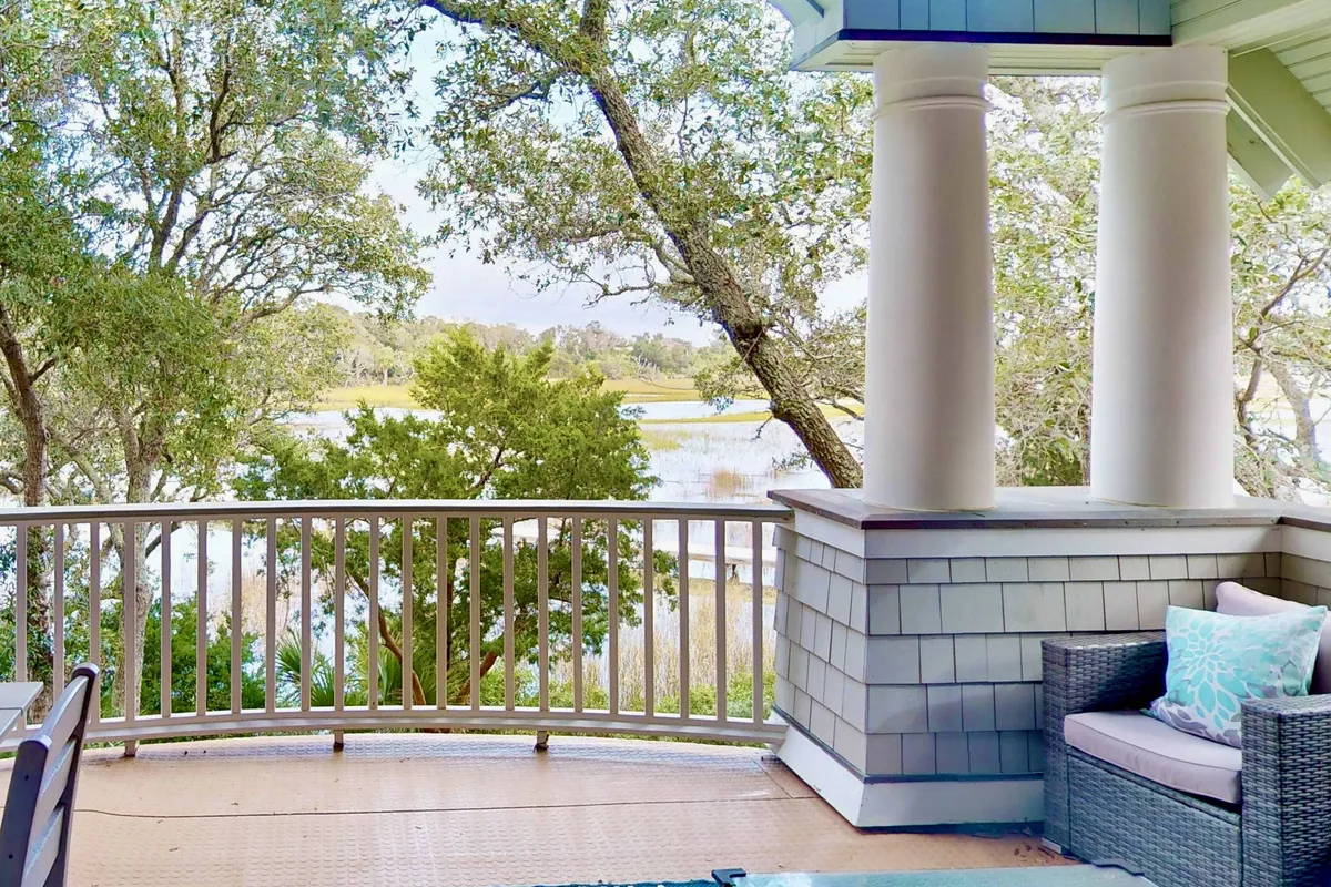 A serene exterior deck area with balcony railing, comfortable outdoor furniture, and a panoramic view of the water and surrounding trees. The space is ideal for relaxing, reading, or gathering as a group to enjoy nature and sunsets.