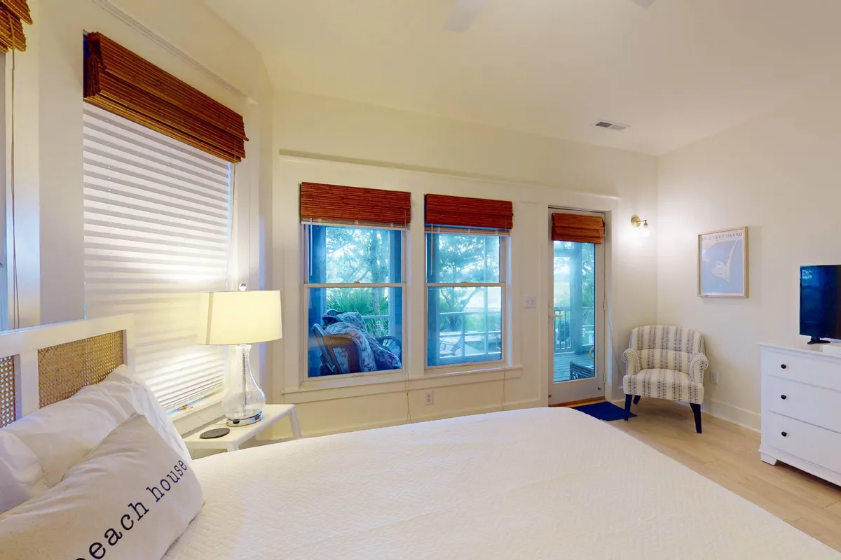 A tranquil bedroom featuring a bed with crisp white linens, large windows that allow for ample natural light, a comfortable chair, and a nightstand with a lamp. The room feels peaceful and inviting, ideal for restful nights or quiet afternoons with a book.