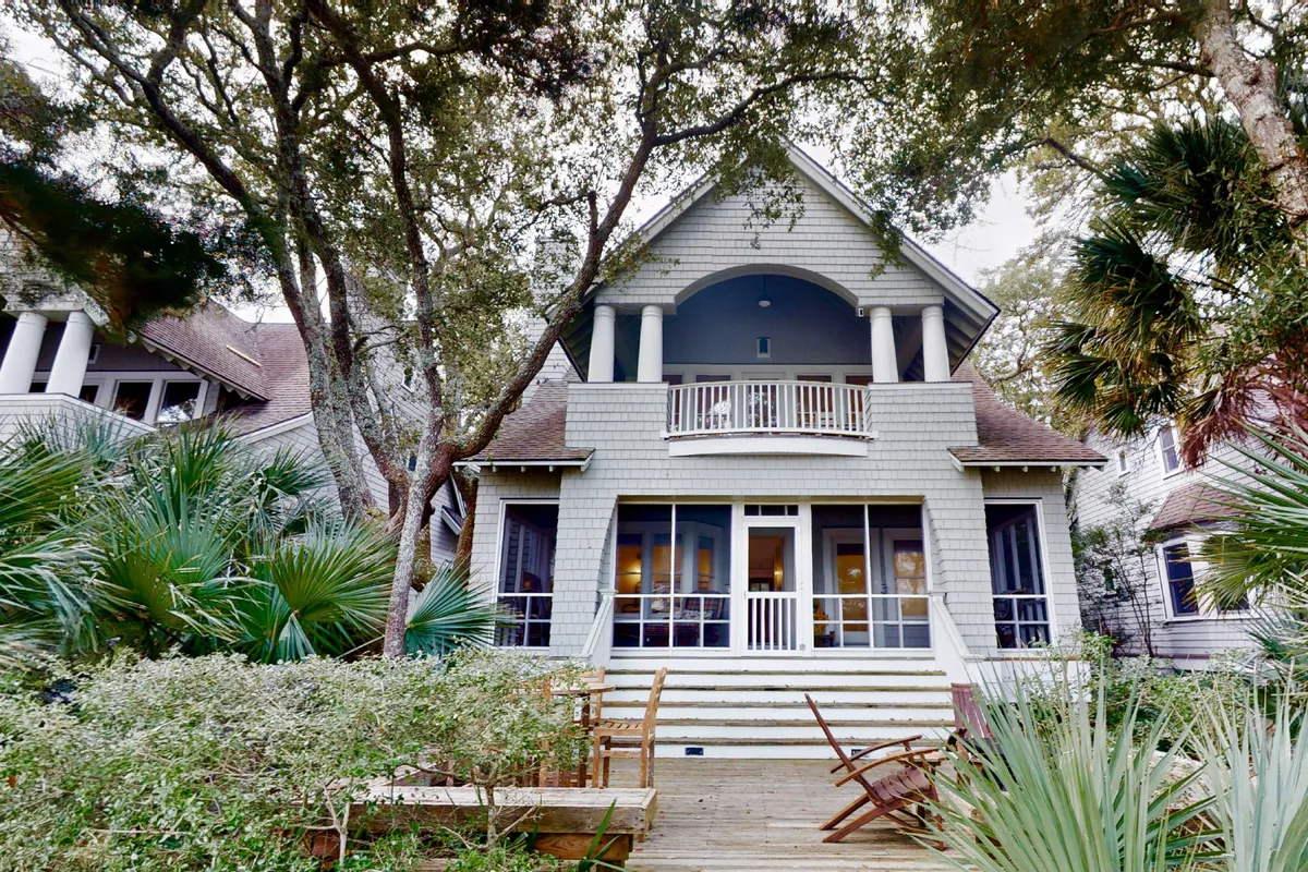 The image shows the grand entrance to Blue Crab Cottage, complete with a welcoming porch featuring columns and surrounded by tranquil greenery. The porch invites relaxation and offers a glimpse of the home's charming exterior.